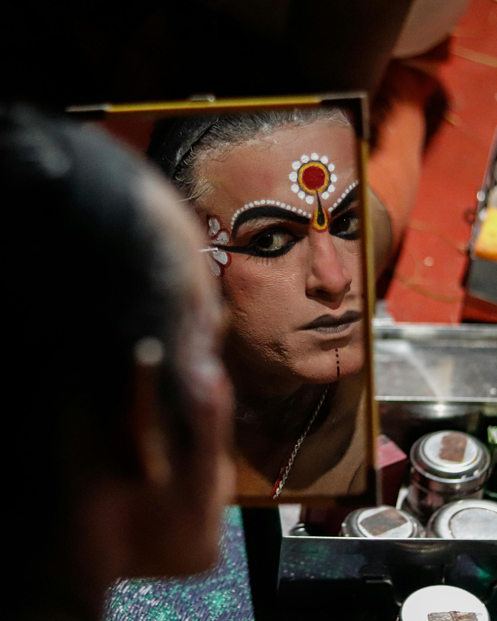 Close-up of Indian artist applying traditional theatre makeup, seen in a mirror reflection.